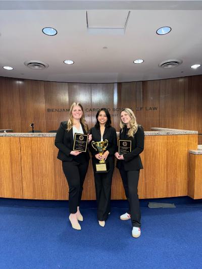 3 students hold up plaques and trophies in a courtroom at Benjamin Cardozo School of Law